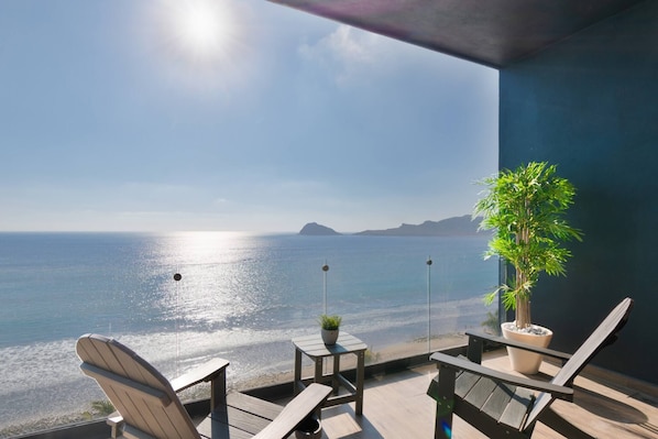 A balcony with a view of the sea, a potted plant, and two wooden chairs.