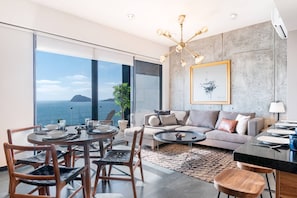 A modern living room with a large window, a sofa, a dining table, and a chandelier.