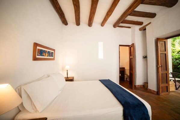 Un dormitorio con cama, mesita de noche, lámpara, cuadro enmarco y puerta de madera que da a un balcón.
