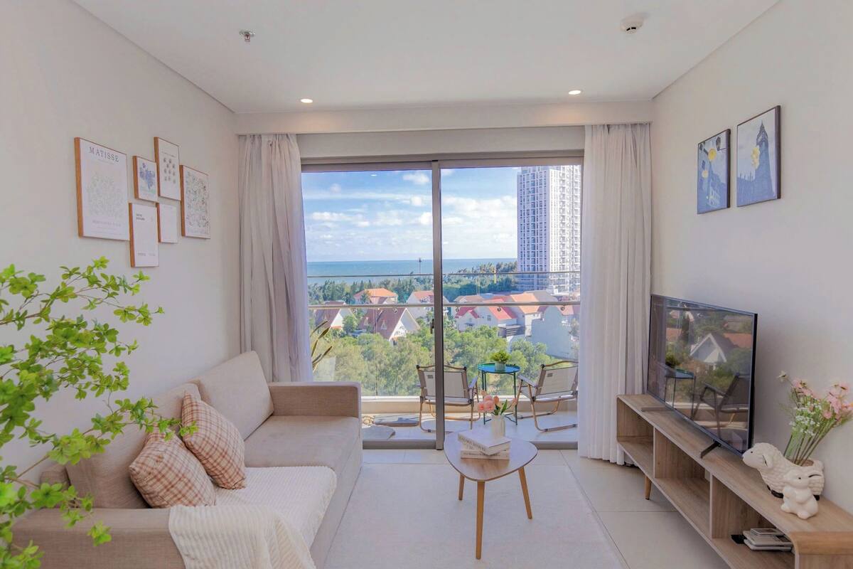 Ein modernes Wohnzimmer mit einer Couch, einem Couchtisch und einem Fernseher auf einem Holzboden. Ein Balkon bietet Blick auf die Stadt und ein Gebäude in der Ferne.