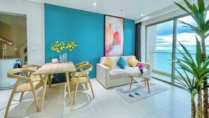 Ein modernes Wohnzimmer mit blauer Akzentwand, einem weißen Sofa mit bunten Kissen, einem Holzesstisch mit Stühlen und einem großen Fenster mit Meerblick.