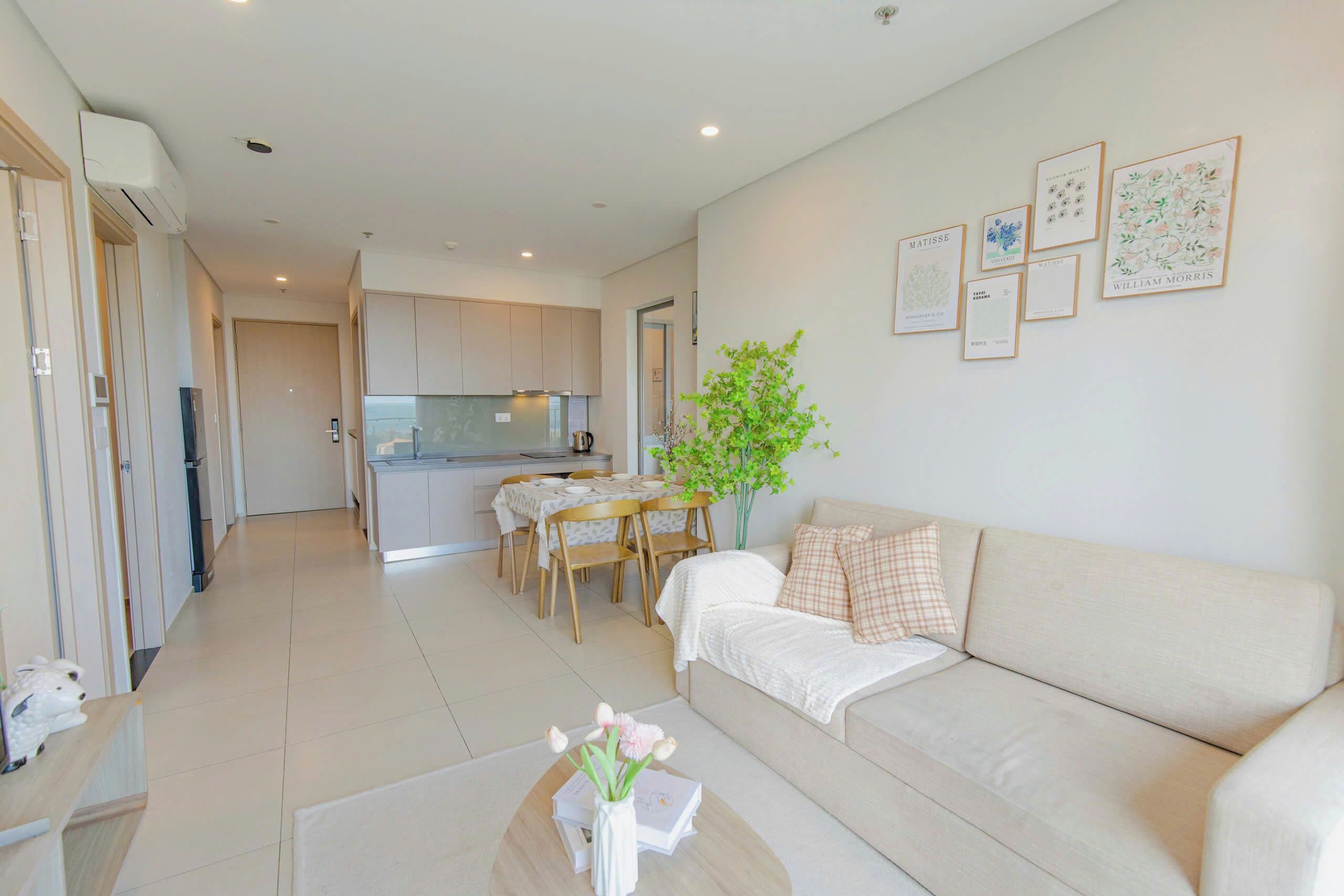 A modern living room with a sofa, a coffee table with flowers, a dining area with a table and chairs, and a kitchen in the background.