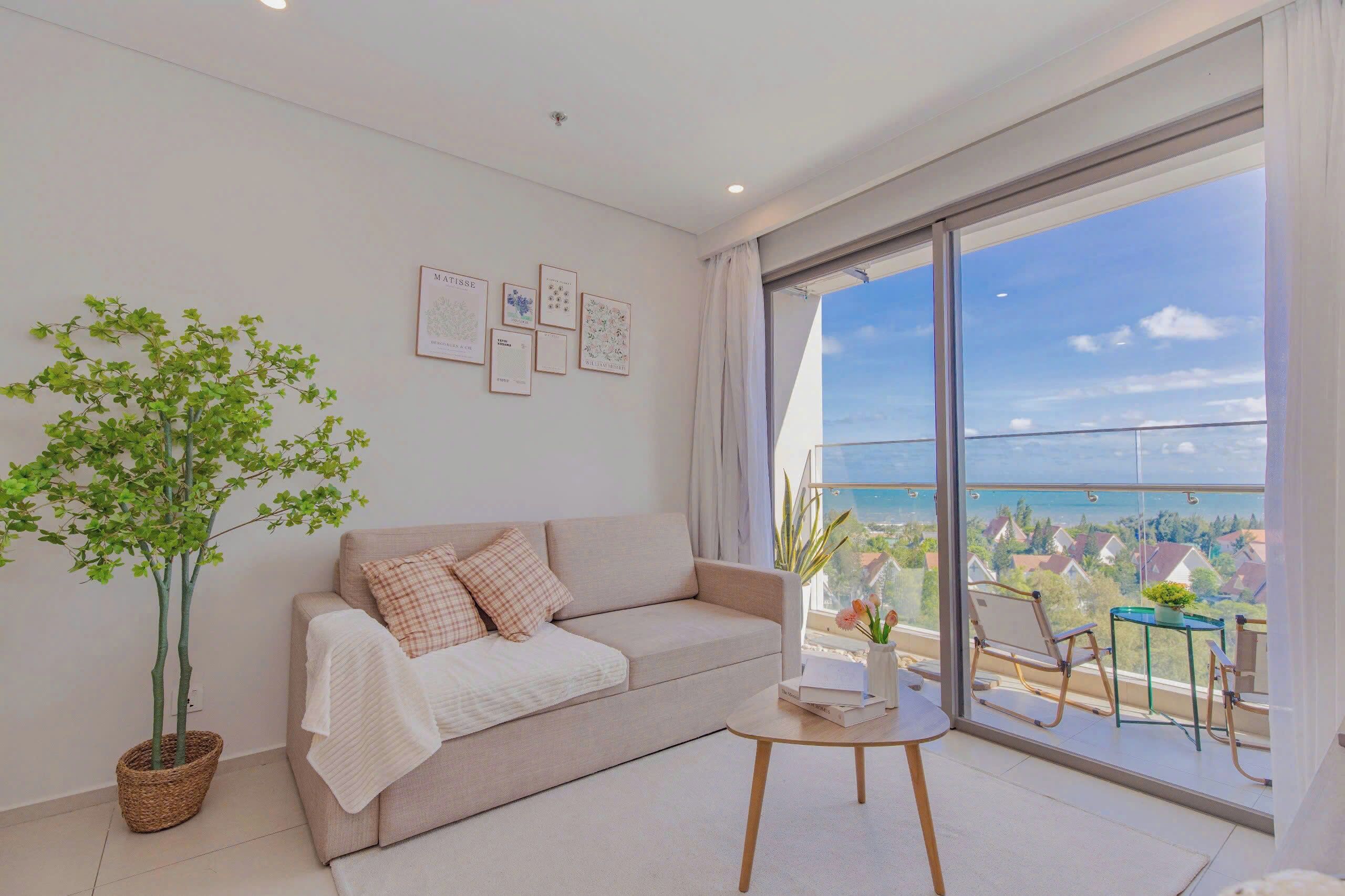 A modern living room with a sofa, a coffee table, a potted plant, and a balcony with a view of the sea.