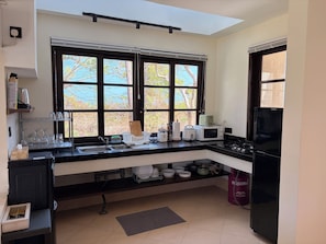 Una cucina con un piano di lavoro nero, un fornello, un forno a microonde e un frigorifero. Si vede una finestra con persiane e una vista su alberi.