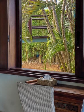 Une fenêtre donnant sur un jardin tropical, une chaise en osier et une petite table en bois agrémentée d’un objet décoratif.
