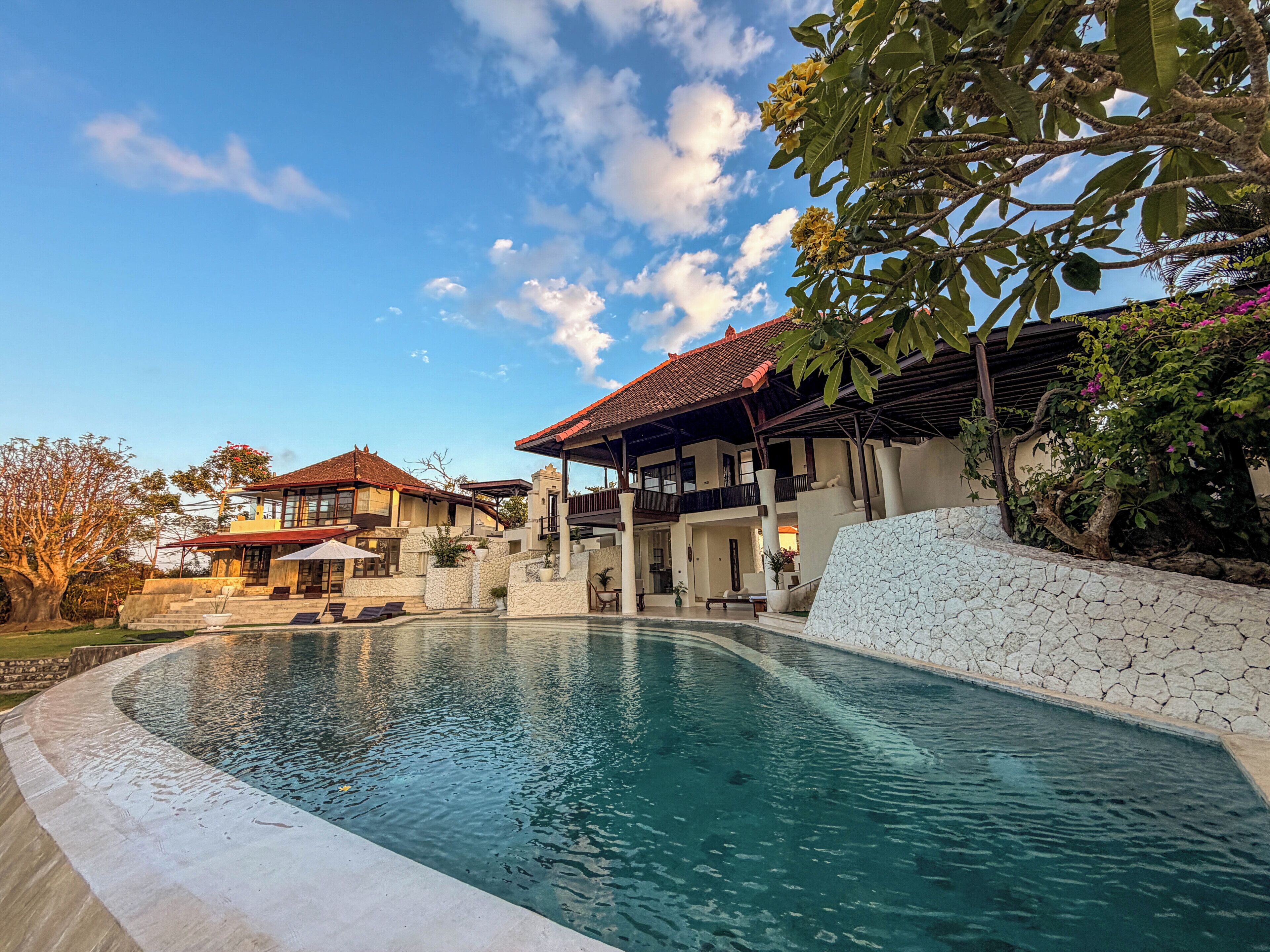 A pool area with a stone wall, a covered seating area, and a building with a red-tiled roof.