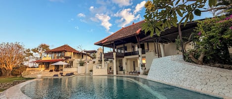 A pool area with a stone wall, a covered seating area, and a building with a red-tiled roof.