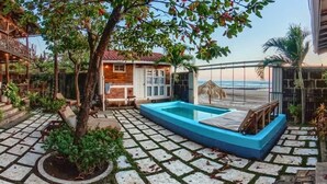 A pool area with a wooden deck, a small building with a glass door, and a view of the ocean.
