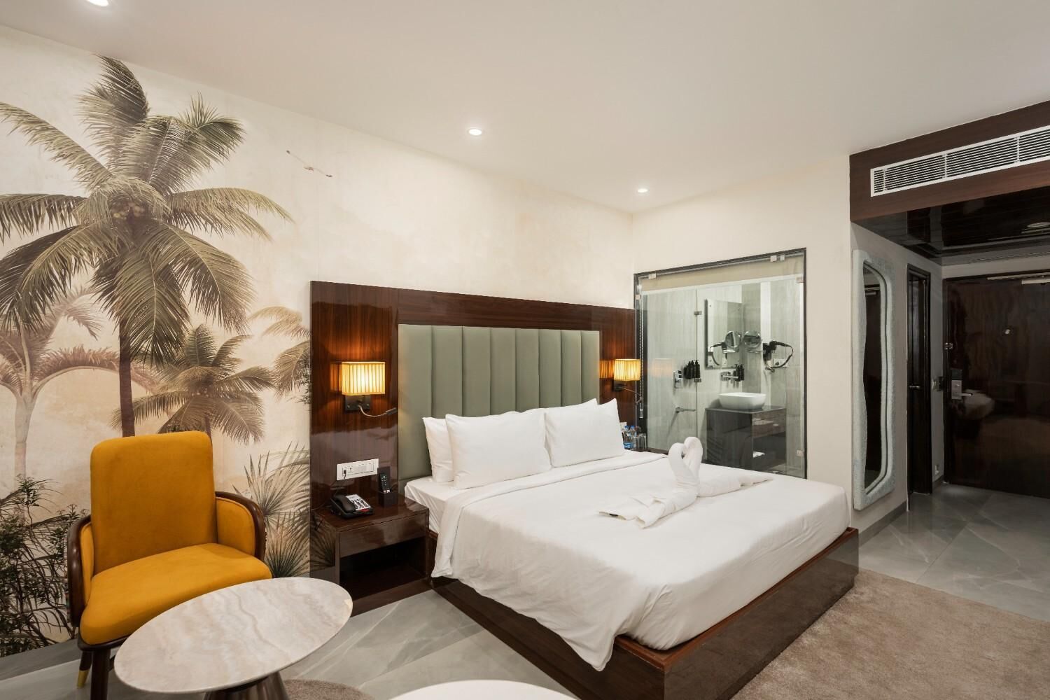 A hotel room with a bed, a nightstand, a chair, a small table, a mirror, and a door leading to a bathroom.