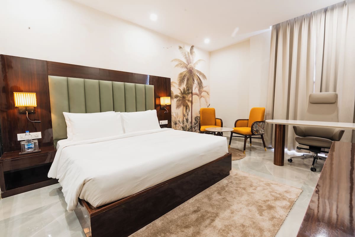 A hotel room with a large bed, a desk, two chairs, a lamp, and a palm tree mural.