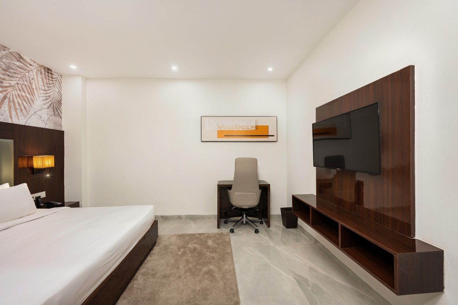 A hotel room with a bed, desk, chair, TV, and artwork on the wall.