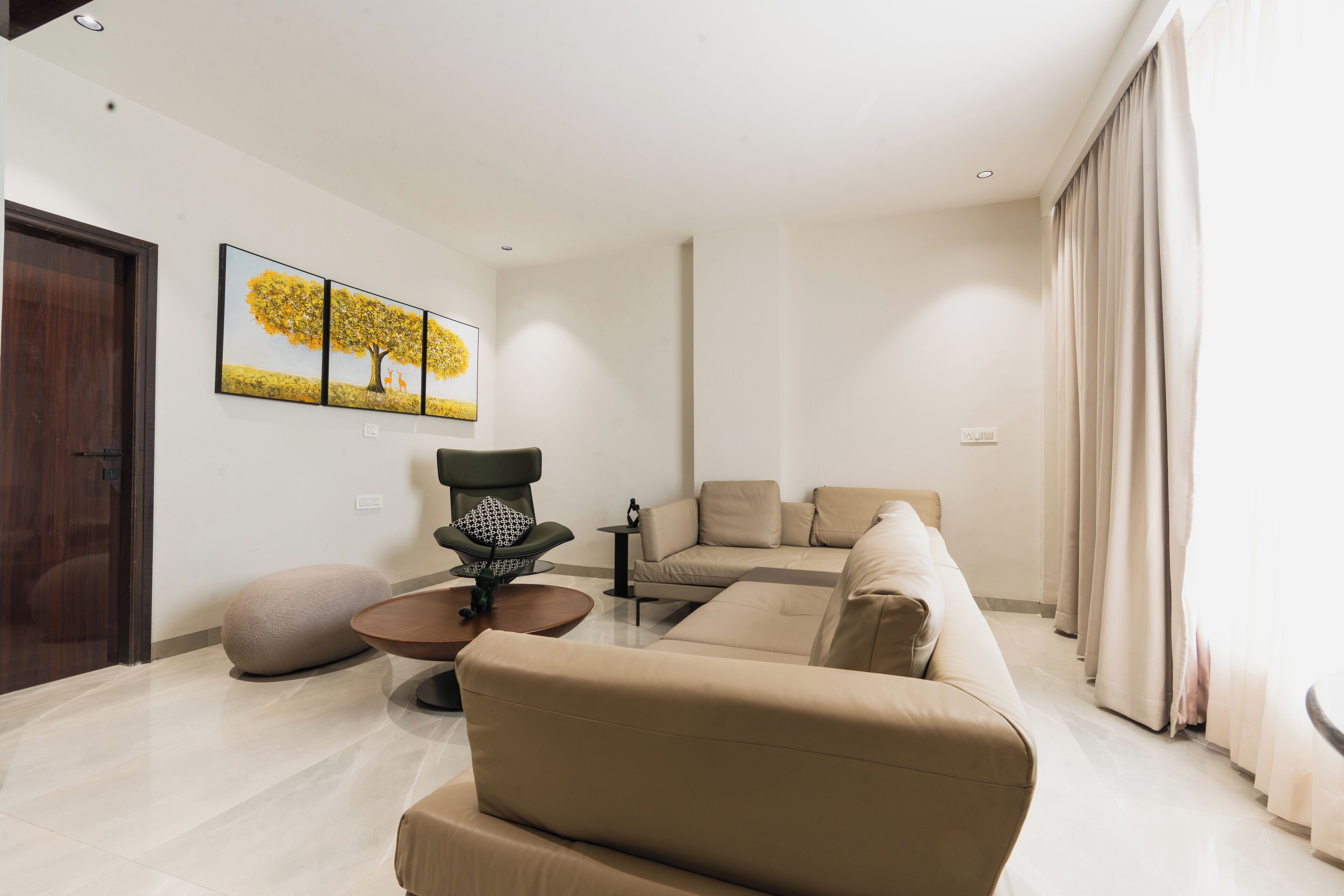 A modern living room with a beige sofa, a round coffee table, and a unique chair. There is a painting on the wall and a door to the left.