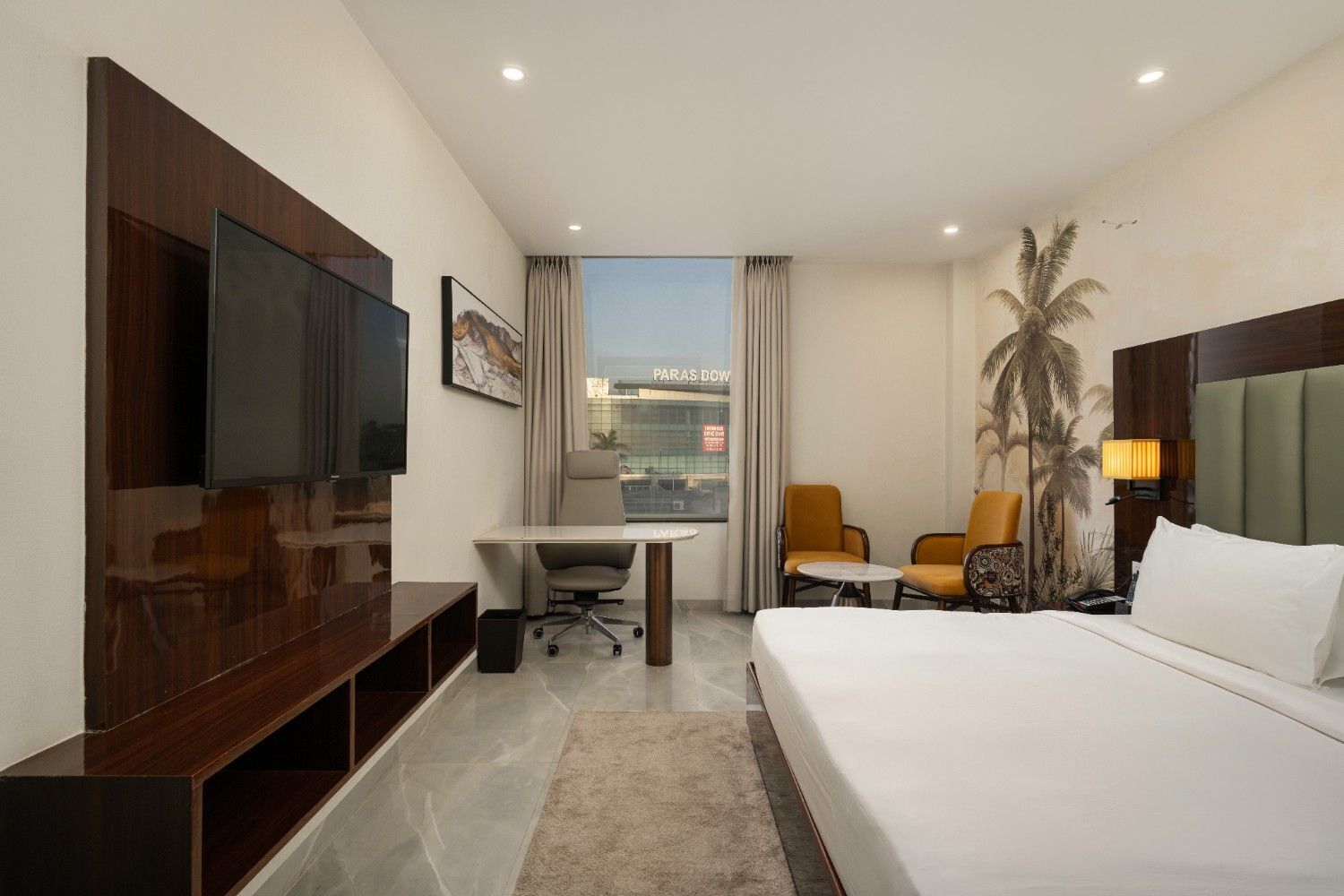 Ein Hotelzimmer mit Bett, Schreibtisch, zwei Stühlen, Fernseher und einem großen Fenster mit Blick auf ein Gebäude.