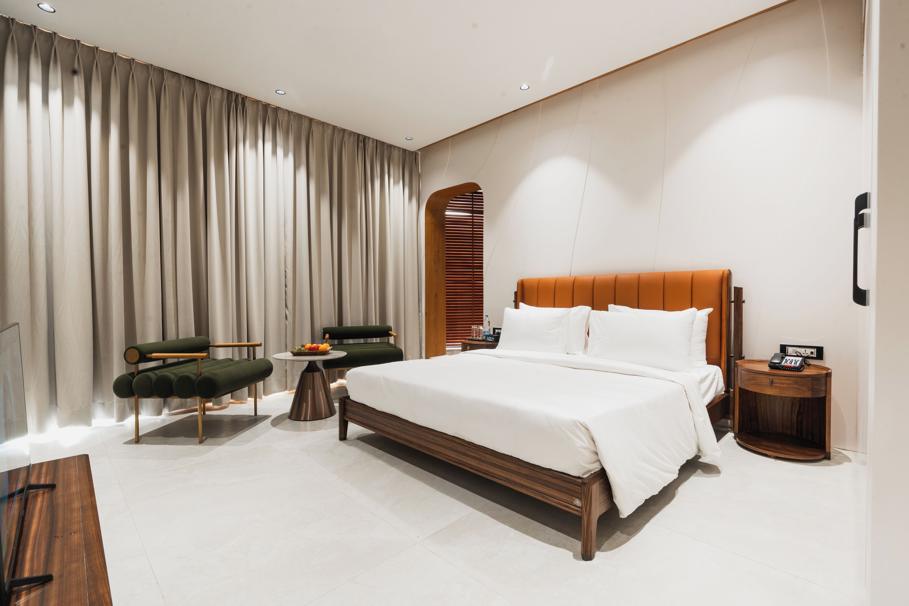 A modern hotel room with a large bed, a seating area with a sofa and armchairs, a small side table, and a wooden nightstand.