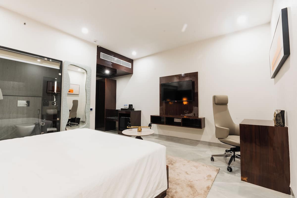 A modern hotel room with a large bed, a desk, a chair, a TV, and a bathroom visible through a glass partition.
