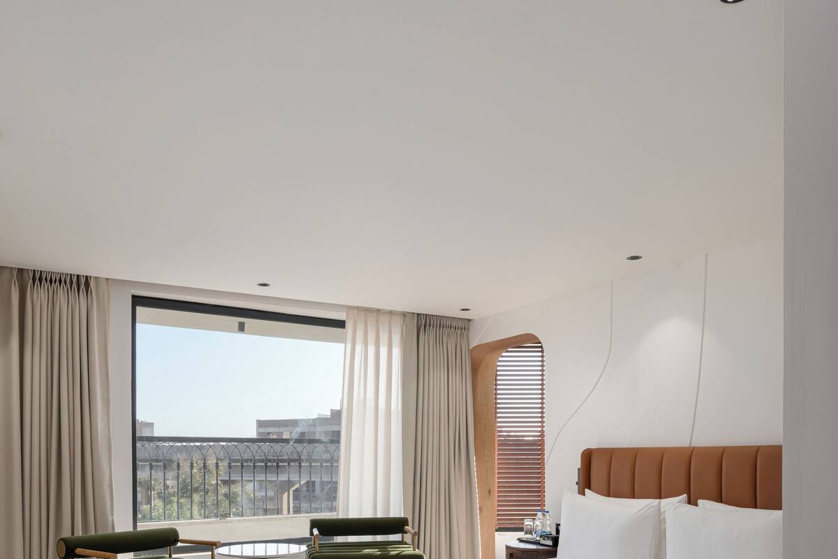A modern hotel room with a large bed, a round rug, a seating area with two chairs, and a view of the city through a large window.
