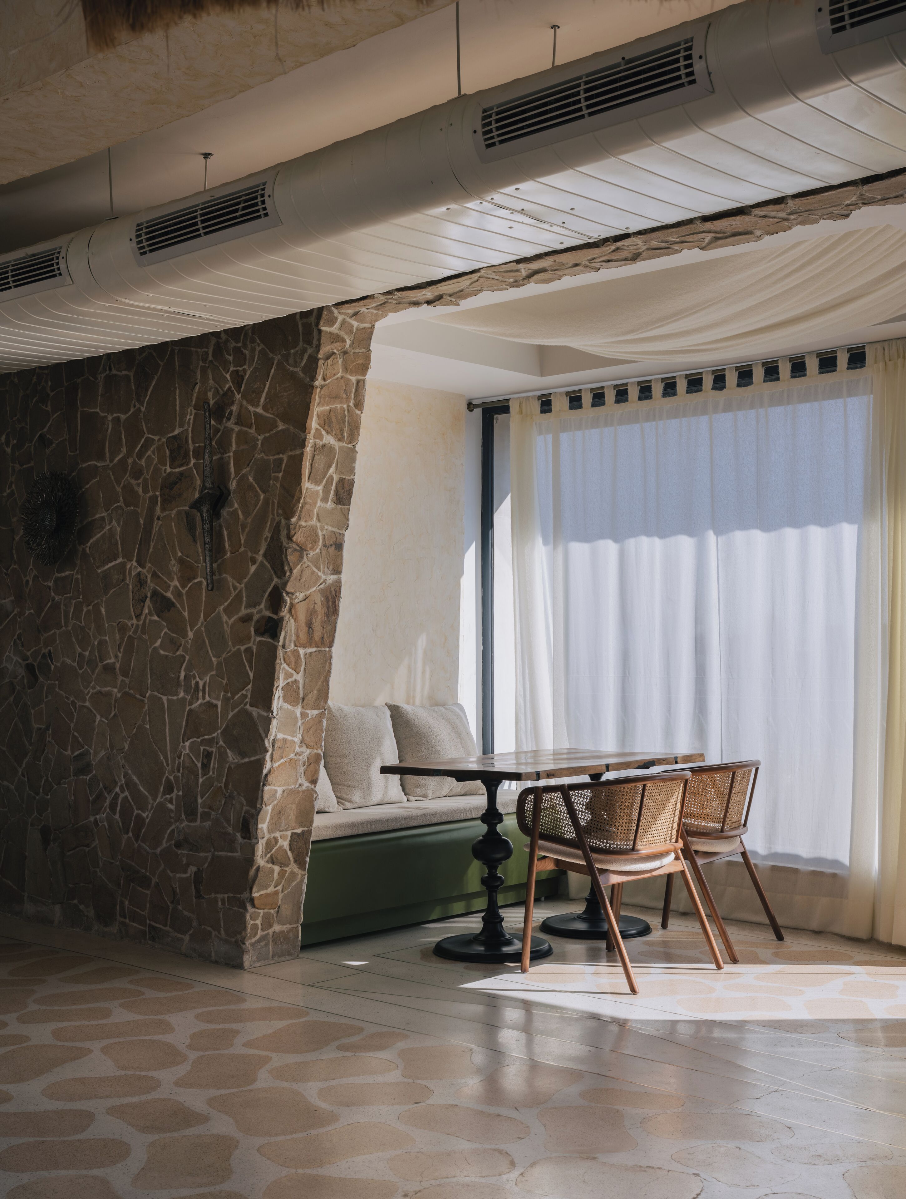 A stone wall, a sofa, a table, and chairs in a room with a large window and curtains.