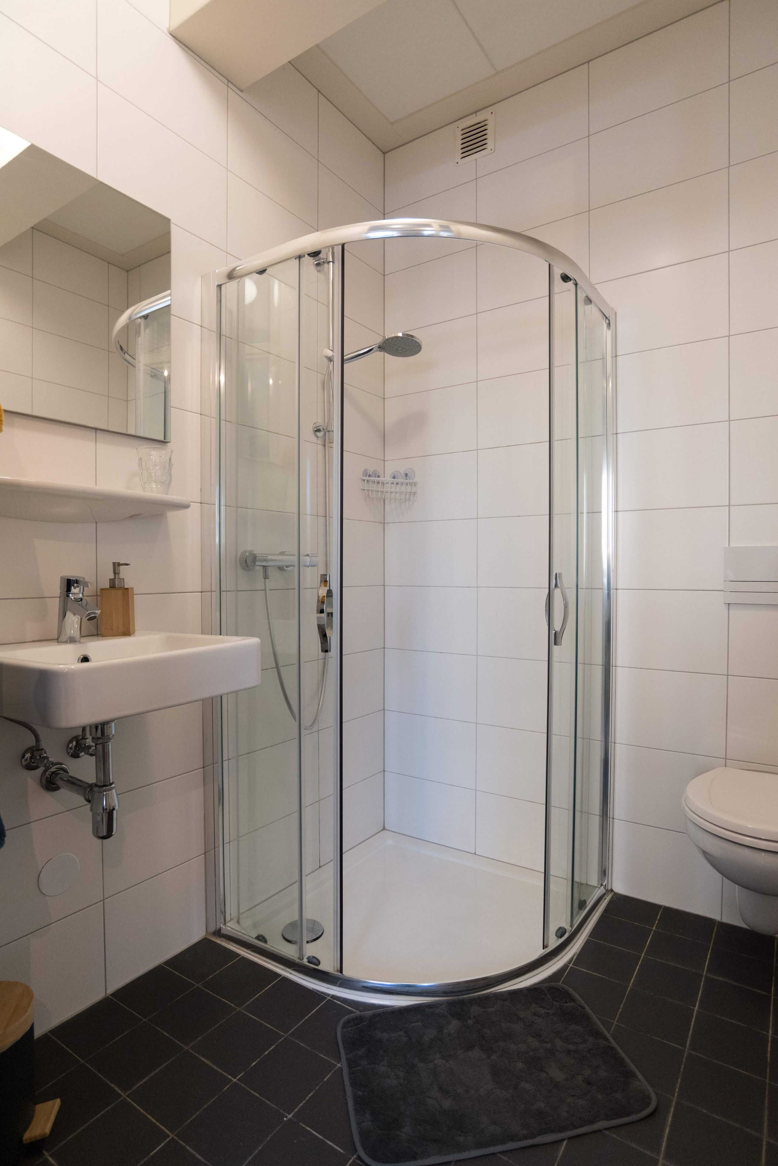 Een douche met glazen wand, een witte wastafel en een toilet in een badkamer met witte betegelde wanden.