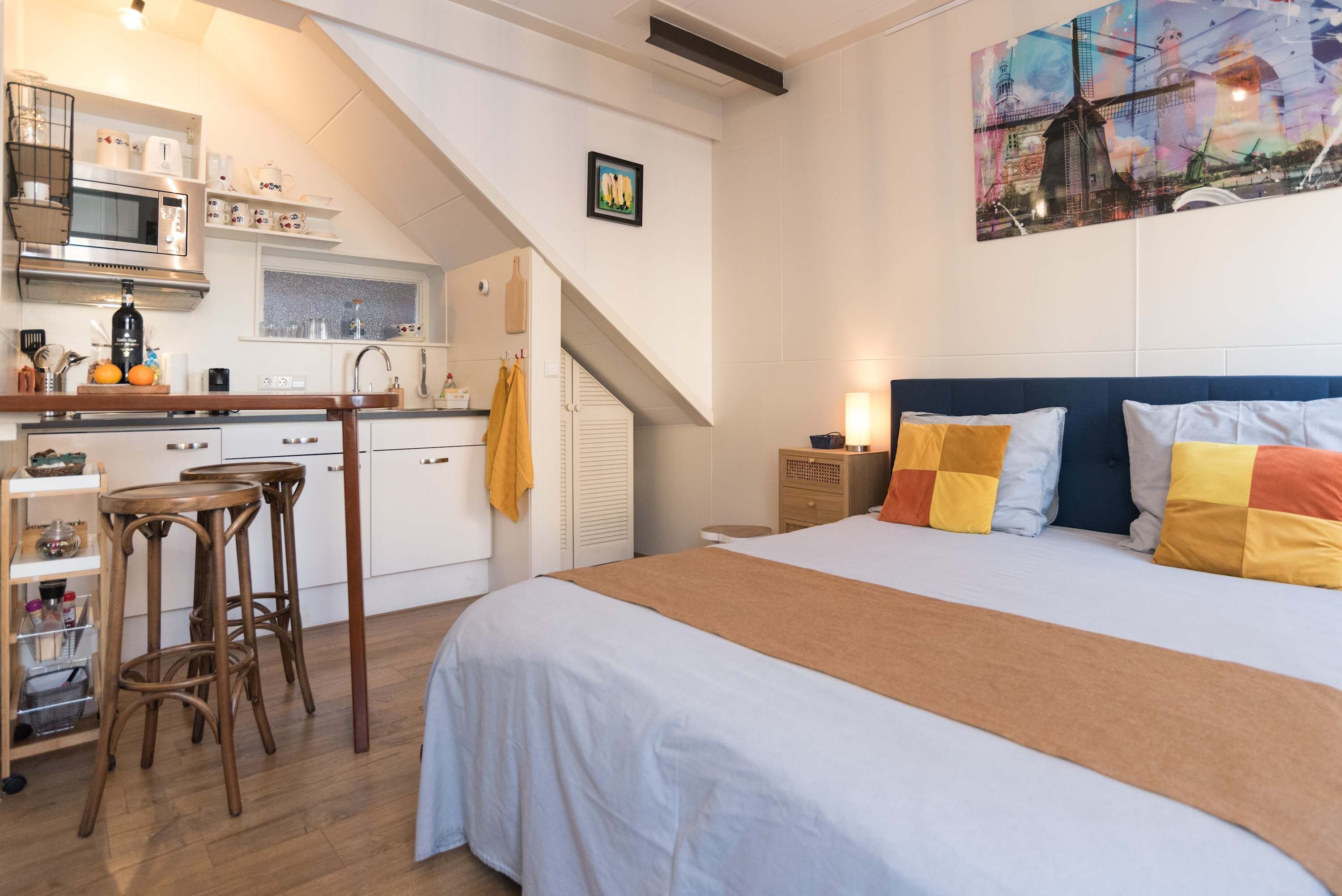 A bedroom with a bed, a nightstand, and a desk with stools. There is a kitchen area with a microwave, a sink, and a stove.