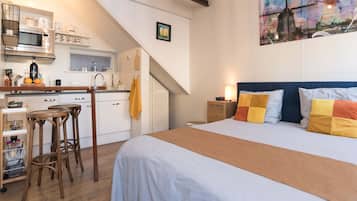 A bedroom with a bed, a nightstand, and a desk with stools. There is a kitchen area with a microwave, a sink, and a stove.