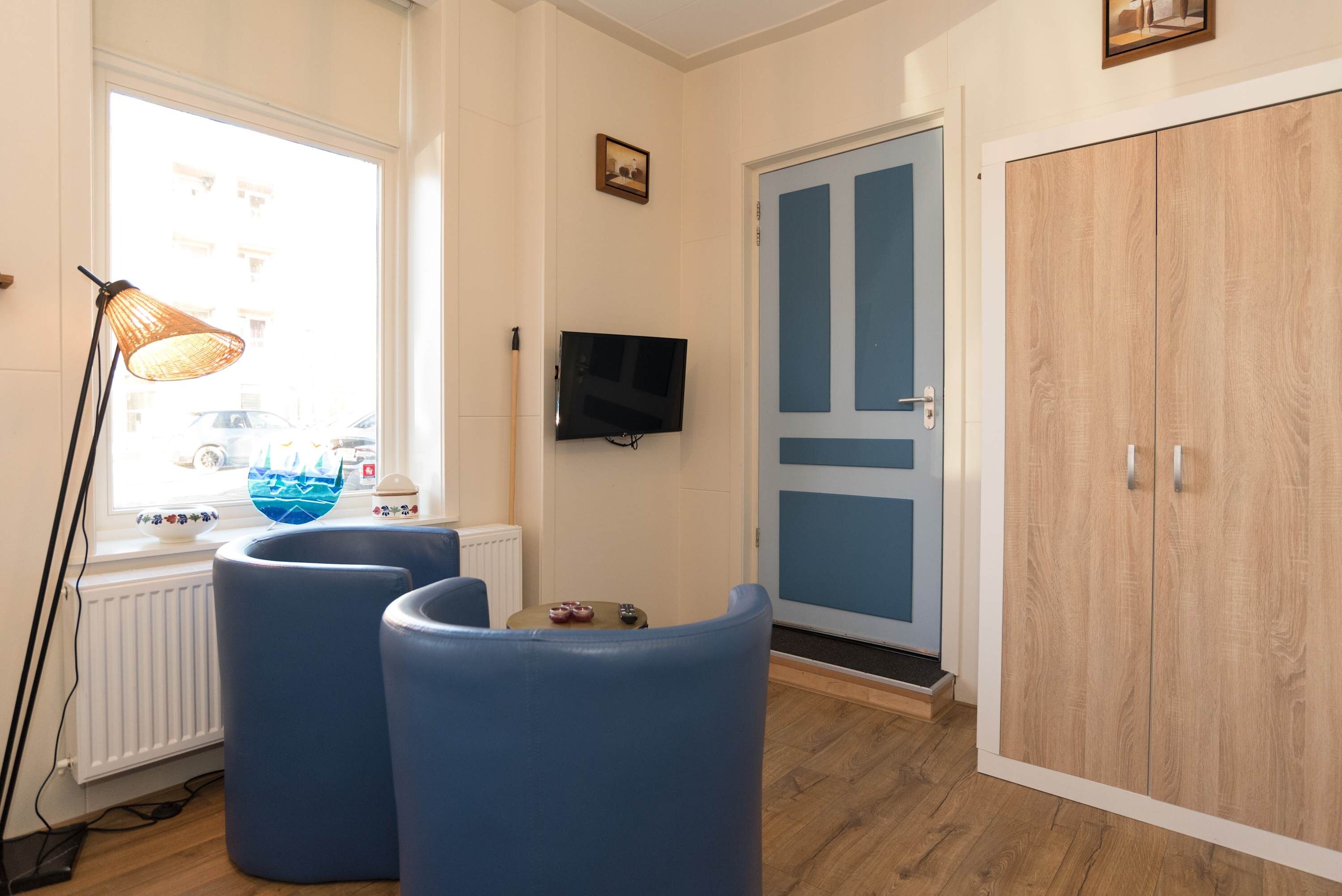 A room with a large window, a television, a wardrobe, and two blue chairs.