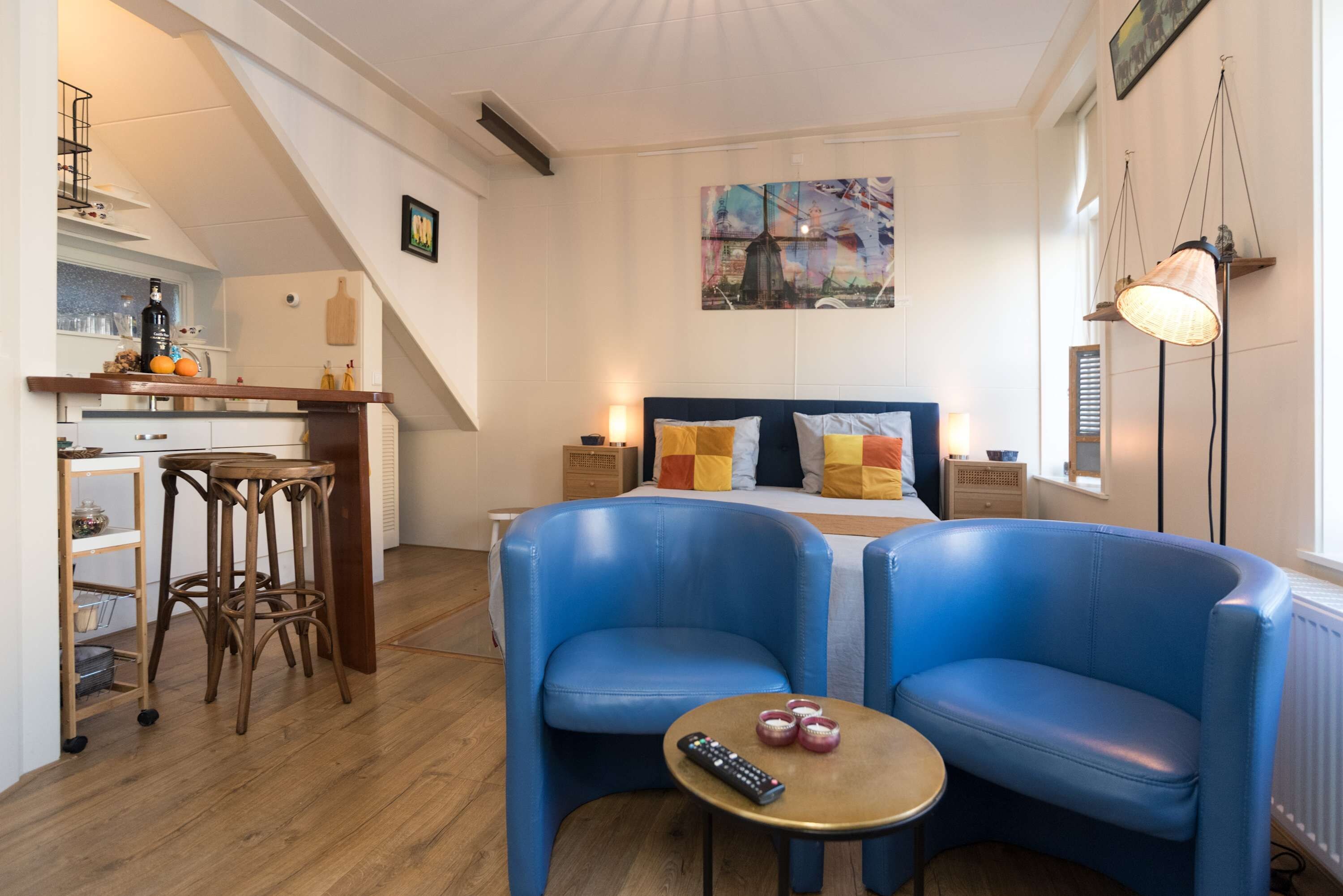 A modern hotel room with a bed, two blue armchairs, a small table with a remote control, and a painting on the wall.
