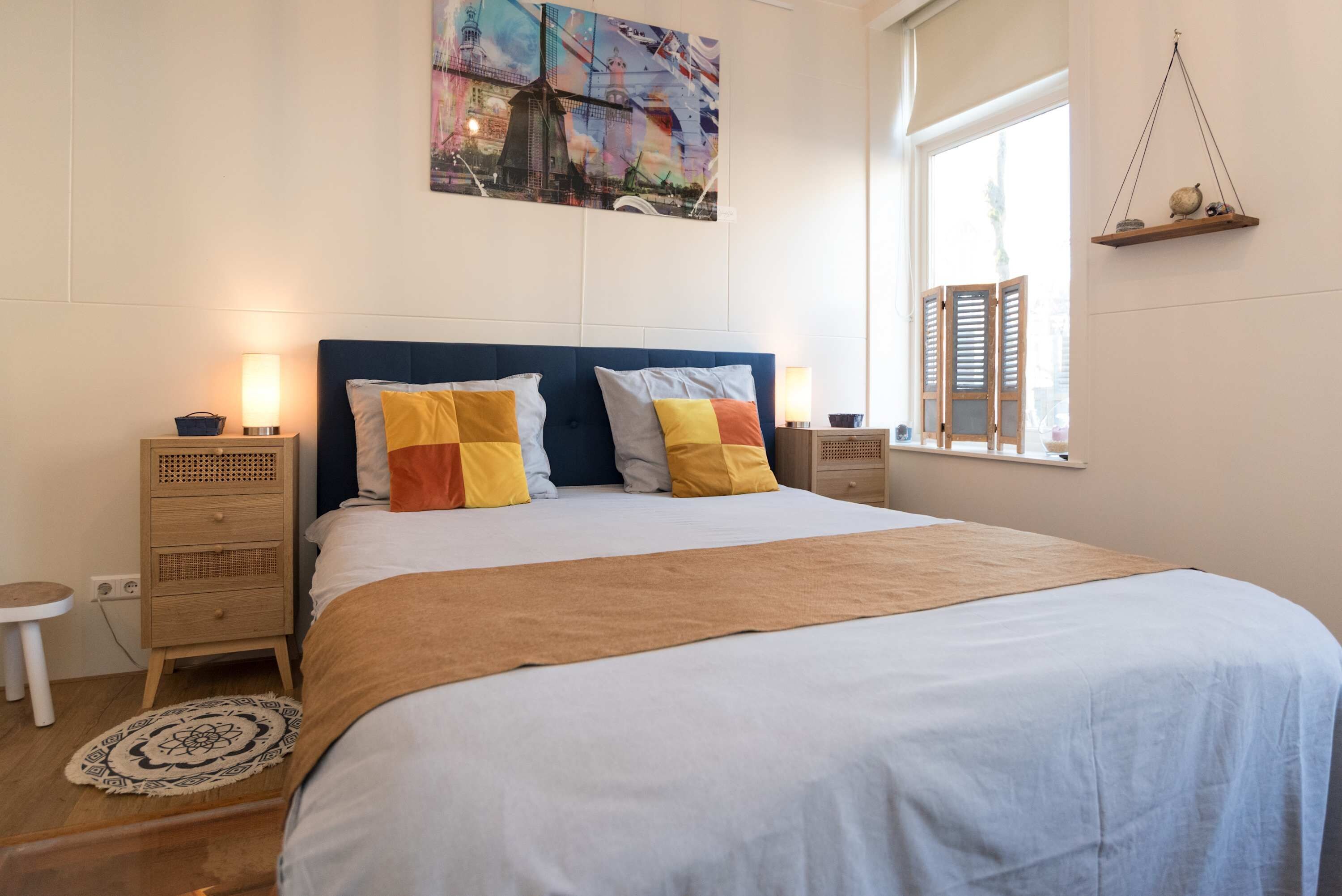 A neatly made bed with a blue headboard, two bedside tables with lamps, a window with shutters, and a wall-mounted shelf with decorative items.
