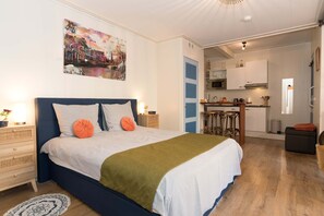 A bedroom with a bed, bedside tables, a nightstand, a painting, a kitchen area with cabinets and a countertop, and a dining area with stools. - Luttik Oudorp 30B Apartment Alkmaar (Alkmaar)