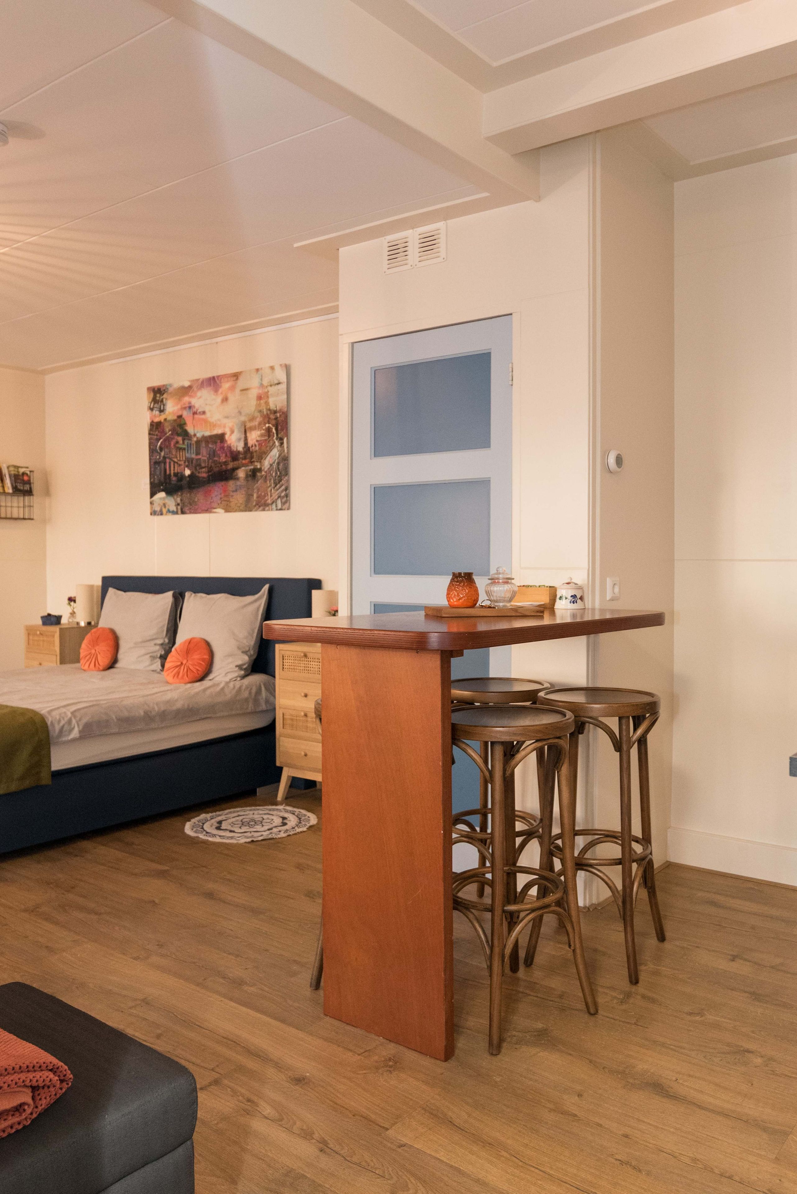 A compact living space with a bed, a small dining area with stools, and a wall-mounted shelf.