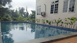 A swimming pool with a tiled edge, surrounded by plants and a building with windows and an air conditioning unit. - Graha Dinar Hotel by HIMA (Cisarua)