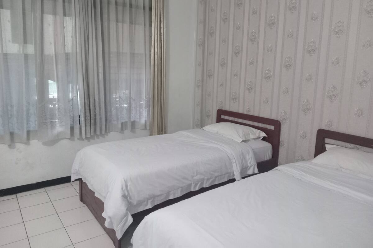 Une chambre avec deux lits simples, une fenêtre avec des rideaux légers et un mur recouvert d’un papier peint à motifs floraux.