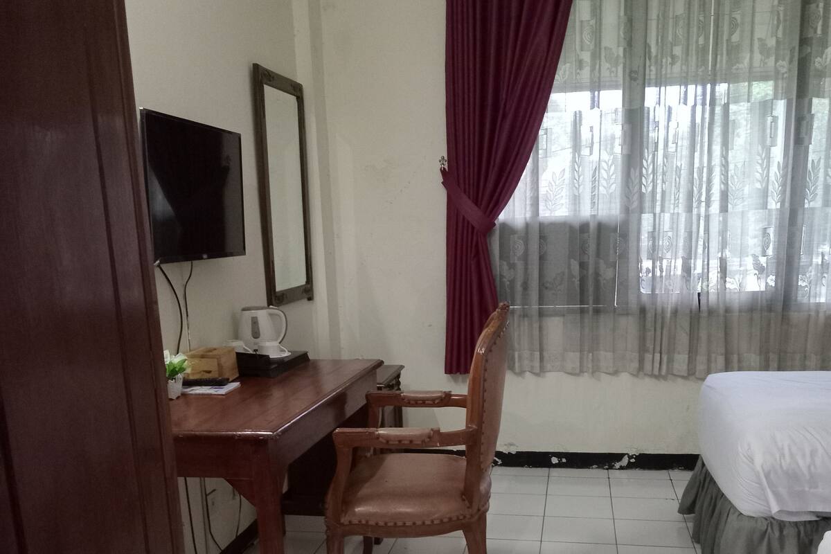 Une chambre d’hôtel avec un bureau en bois, une chaise, un lit, une télévision et une fenêtre avec des rideaux.
