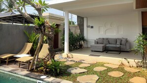 Un'area esterna moderna con piscina, poltrona prendisole e un divano grigio sotto un pergolato bianco.