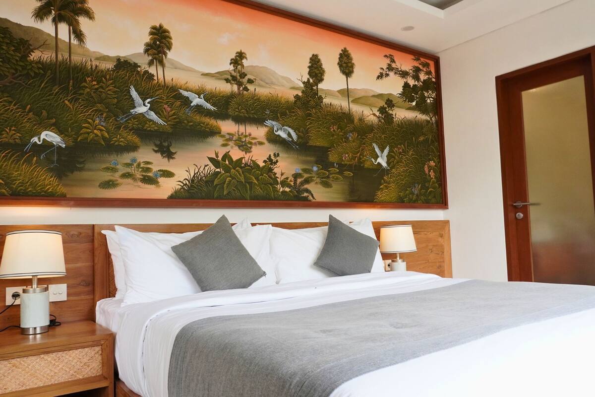 A hotel room with a large bed, two bedside tables with lamps, and a large wall painting of a landscape with birds.