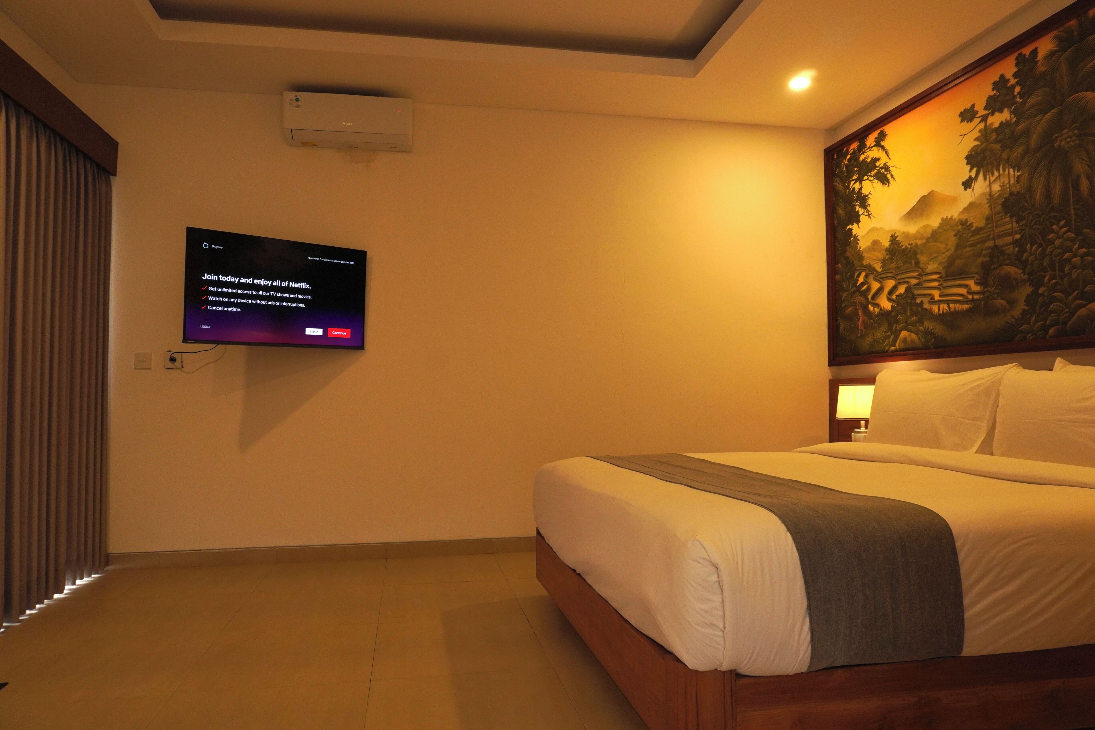 A hotel room with a bed, a mounted TV displaying a Netflix sign, a painting on the wall, and a ceiling light.
