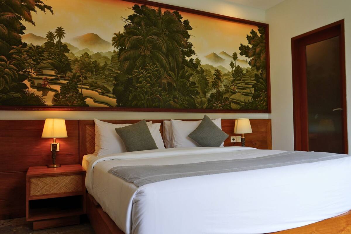 A hotel room with a large bed, two bedside lamps, a wooden headboard, and a large painting of a tropical landscape on the wall.