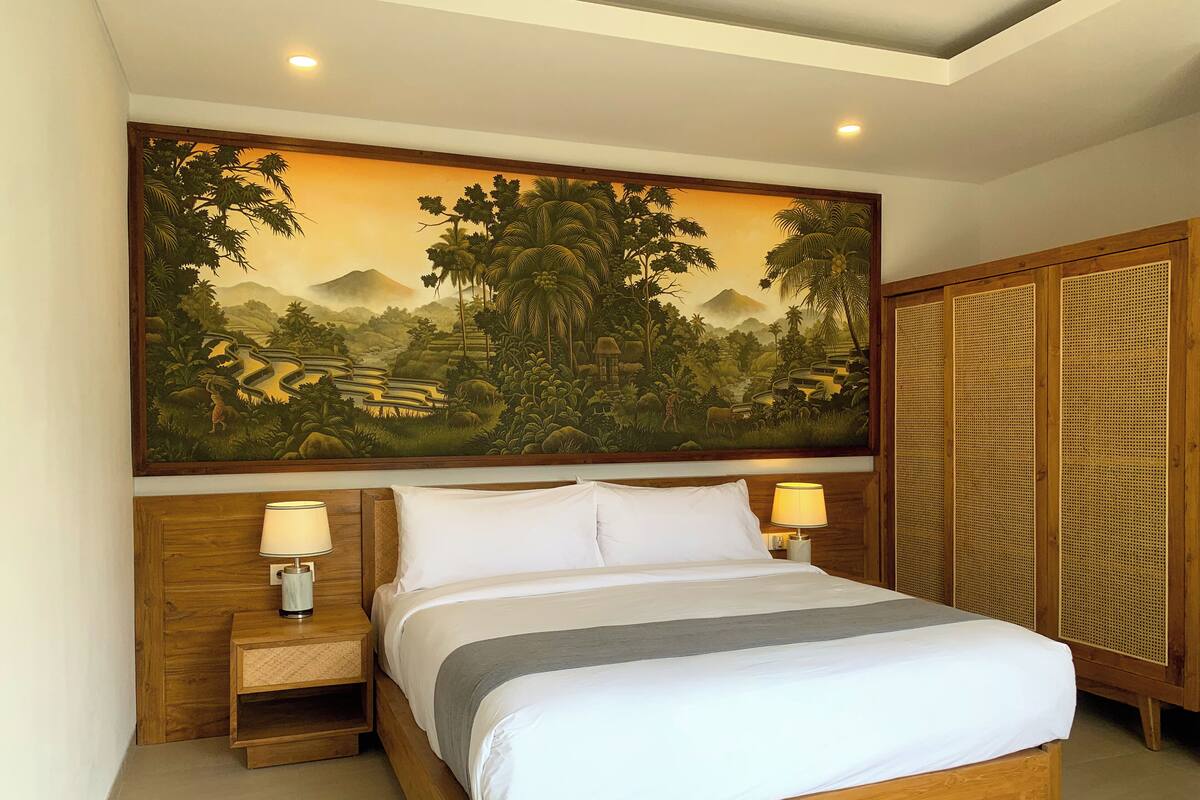 A neatly made bed with white linens, two bedside lamps, and a large painting of a tropical landscape above it.