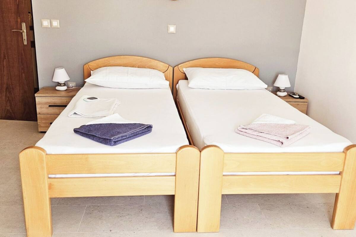 A double room with two wooden beds, each with a nightstand, a lamp, and a folded towel.