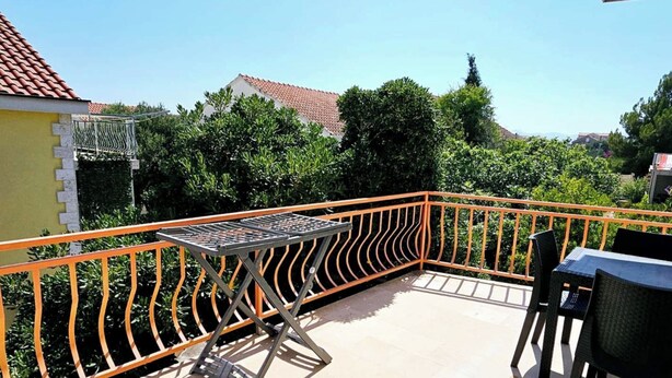 Un balcon doté d’une table et de chaises en métal, entouré de rampes orange, donnant sur un quartier résidentiel avec des arbres et des maisons.