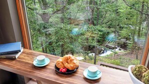 Uma mesa de madeira com um croissant, um prato com framboesas e duas chávenas, com vista para uma área arborizada e um riacho.