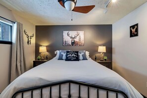 A bedroom with a bed, two bedside tables with lamps, a ceiling fan, a window with curtains, and wall art including a deer head and a mountain scene. - Waterfall Views! Hot Tub, Game Room, Dog Friendly (Bushkill)