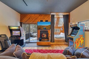 A cozy cabin interior with a fireplace, a gaming console, and a view of the outdoors. - Waterfall Views! Hot Tub, Game Room, Dog Friendly (Bushkill)