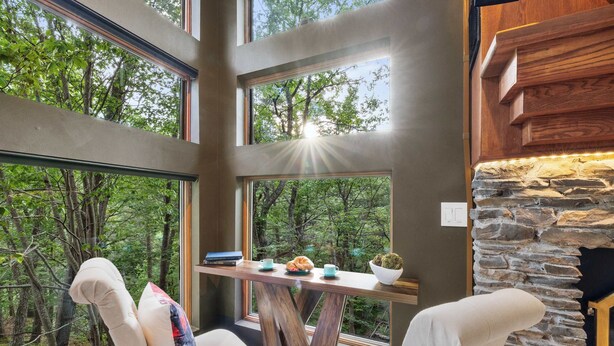 Interior moderno com lareira de pedra, amplas janelas com vista para uma floresta e mesa de madeira com um livro e um croissant.