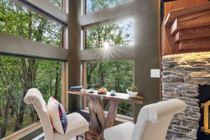 Un interior moderno con chimenea de piedra, ventanas amplias con vista al bosque, y una mesa de madera con un libro y un croissant.
