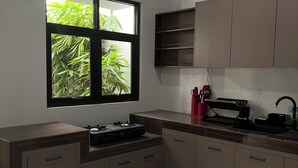 A modern kitchen with a large window, wooden cabinets, a white countertop, and a dining area with a table and chairs. - Aruna Kalasan (Depok)