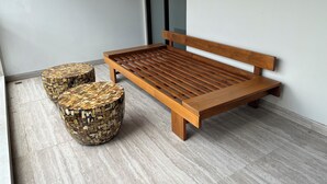 A wooden bench with a slatted seat and backrest, accompanied by two round stools with woven seats, placed on a tiled floor against a plain wall. - Aruna Kalasan (Depok)