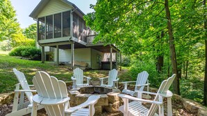 Une maison à deux étages dotée d’une terrasse couverte et d’un foyer extérieur, le tout entouré de chaises Adirondack blanches.