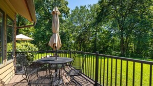 Eine Terrasse mit rundem Tisch und Stühlen, einem Sonnenschirm und Blick auf eine grüne Rasenfläche und Bäume.