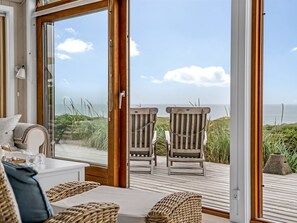 Een kamer met uitzicht op zee, met een houten terras, twee houten stoelen, een witte bank en rieten manden.