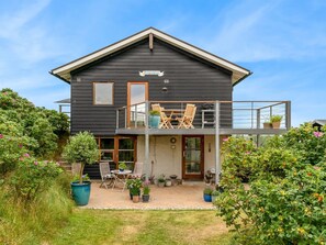 Una casa de dos pisos con un balcón, un pequeño patio con muebles de exterior y varias plantas en macetas.